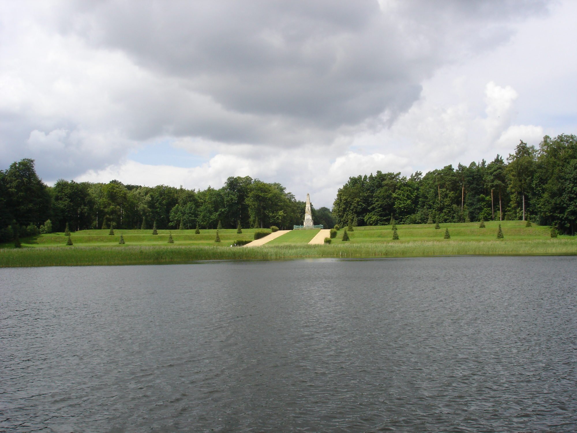 Der Rheinsberger Obelisk ist in der Bildmitte zu sehen. Im Bildvordergrund ist der Grienericksee. Links und rechts stehen Wälder. Fontane nennt den Obelisken die "größte Sehenswürdigkeit" in Rheinsberg.