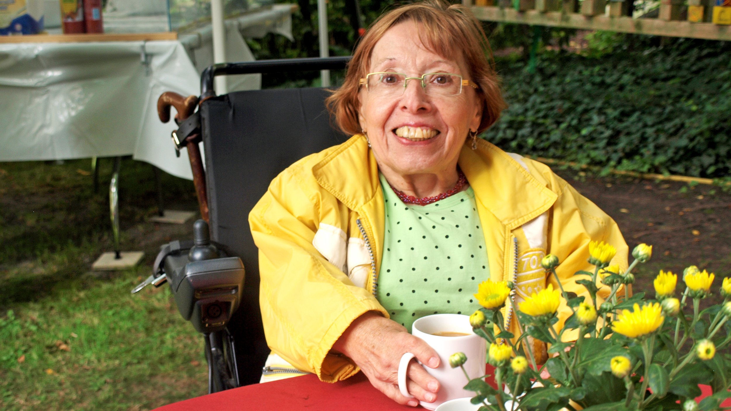 Foto: Ingrid Koch in gelber Windjacke mit ihrem E-Rollstuhl im Garten sitzend, vor ihr ein Kaffeebecher und ein gelber Strauß Blumen