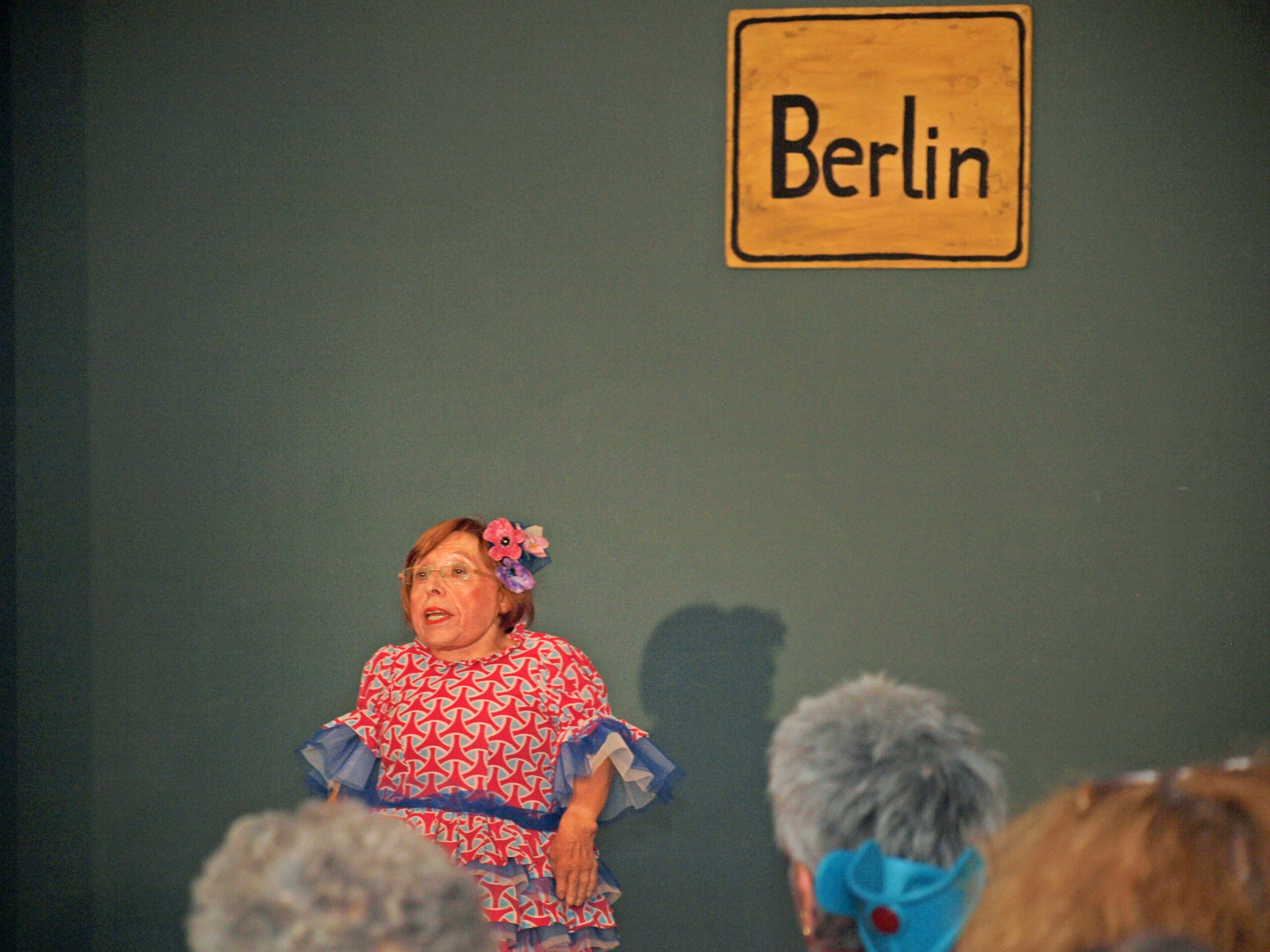 Foto: Ingrid Koch in buntem Kostüm auf der Bühne der Villa Donnersmarck, im Hintergund ein handgemaltes Ortsschild mit der Aufschrift Berlin