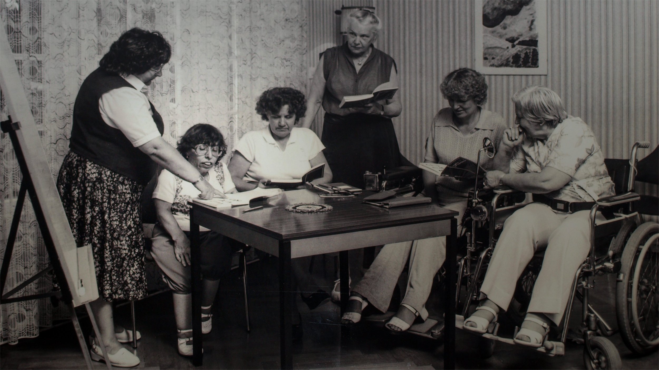 Foto in Schwarz-Weiß: Ingrid Koch als junge Frau am Tisch mit drei weiteren weiblichen Gästen, stehend zwei Mitarbeiterinnen der Freizeitstätte; die Szenerie ist historisch