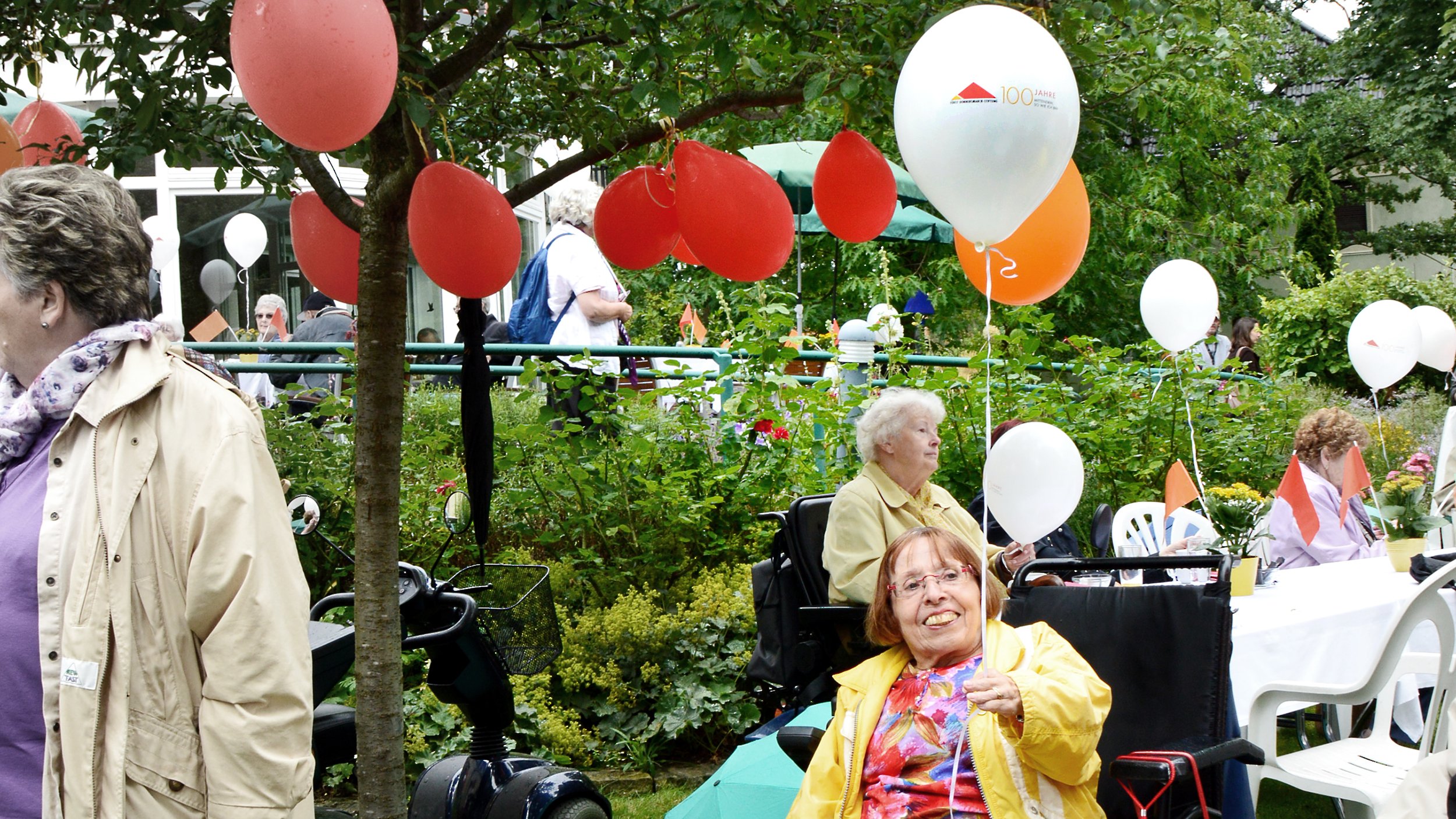 Foto: Ingrid Koch im vollen Garten der Villa Donnersmarck, sie hält einen weißen Gasballon mit der Aufschrift: "100 Jahre Fürst Donnersmarck-Stiftung"