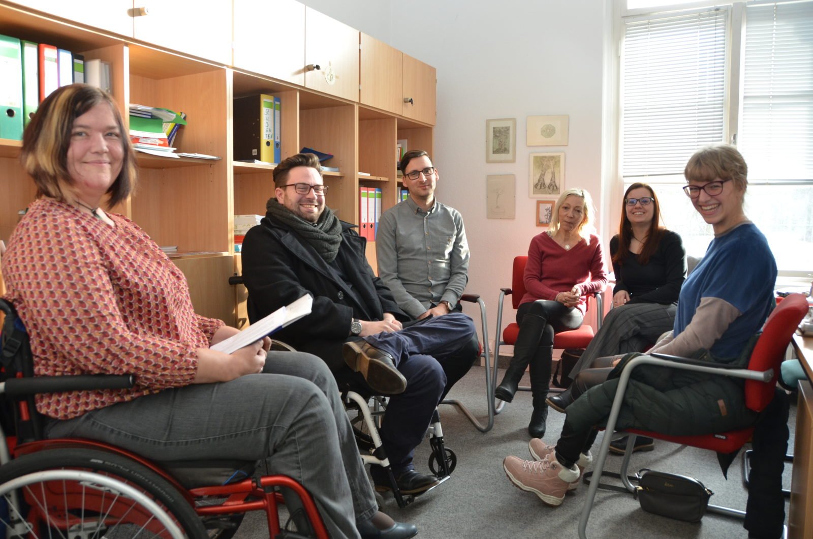 Eine Gruppe Menschen sitzt im Gesprächskreis. Links eine junge Frau und ein junger Mann im Rollstuhl.