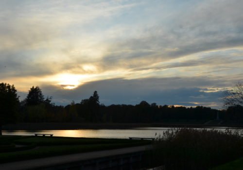 Ein Sonnenuntergang bei wolkenbedecktem Himmel, direkt am Wasser. Über den See hinweg sind im Hintergrund sind ein Wald und ein Obelisk.