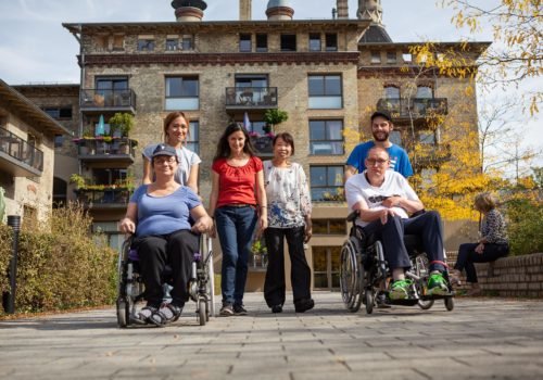 Eine Gruppe Menschen, eine Rollstuhlfahrerin und ein Rollstuhlfahrer, sowie drei Frauen und ein Mann zu Fuß, vor einem alten Backsteingebäude mit Balkons auf denen Pflanzen und Möbel stehen.