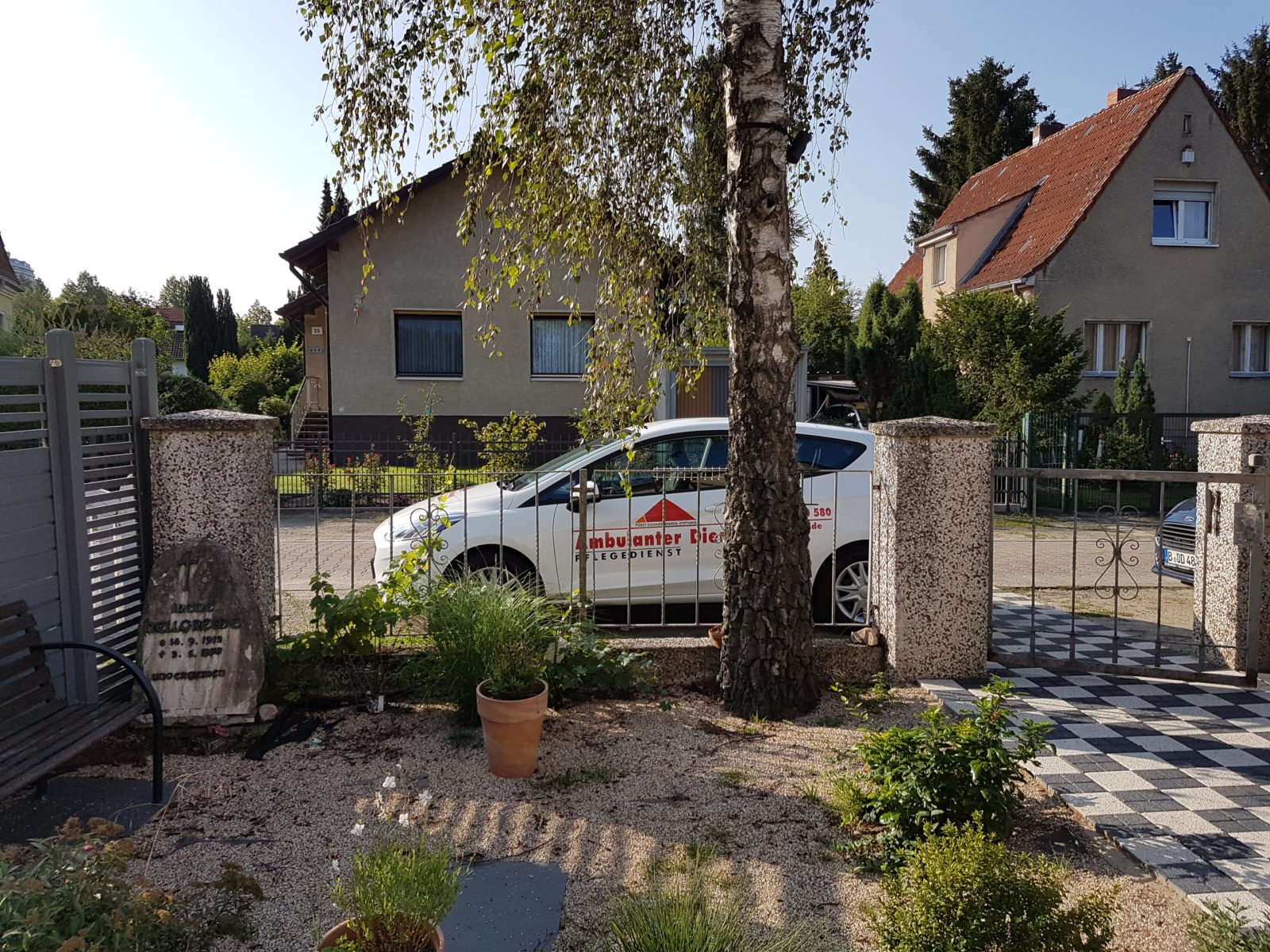 Ein Auto des Ambulanten Dienstes, mit Logos beklebt, steht in einem Wohngebiet am Straßenrand. Das Bild ist aus einem Vorgarten heraus fotografiert. Das Auto steht hinter einem Zaun, davor ein Baum und mehrere Töpfe mit Pflanzen.