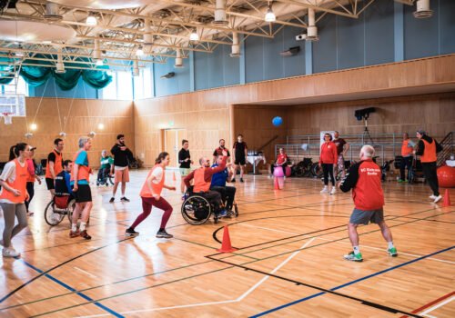 Menschen mit und ohne Behinderung, manche im Rollstuhl, spielen gemeinsam eine Ballsportart in einer Turnhalle.