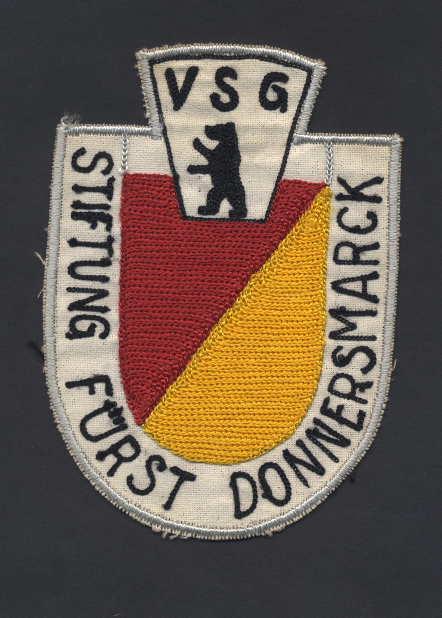 Das Abzeichen der VSG Donnersmarck: Ein Rot-Gelbes Wappen, darüber der Berliner Bär und die Beschriftung VSG Stiftung Fürst Donnersmarck.