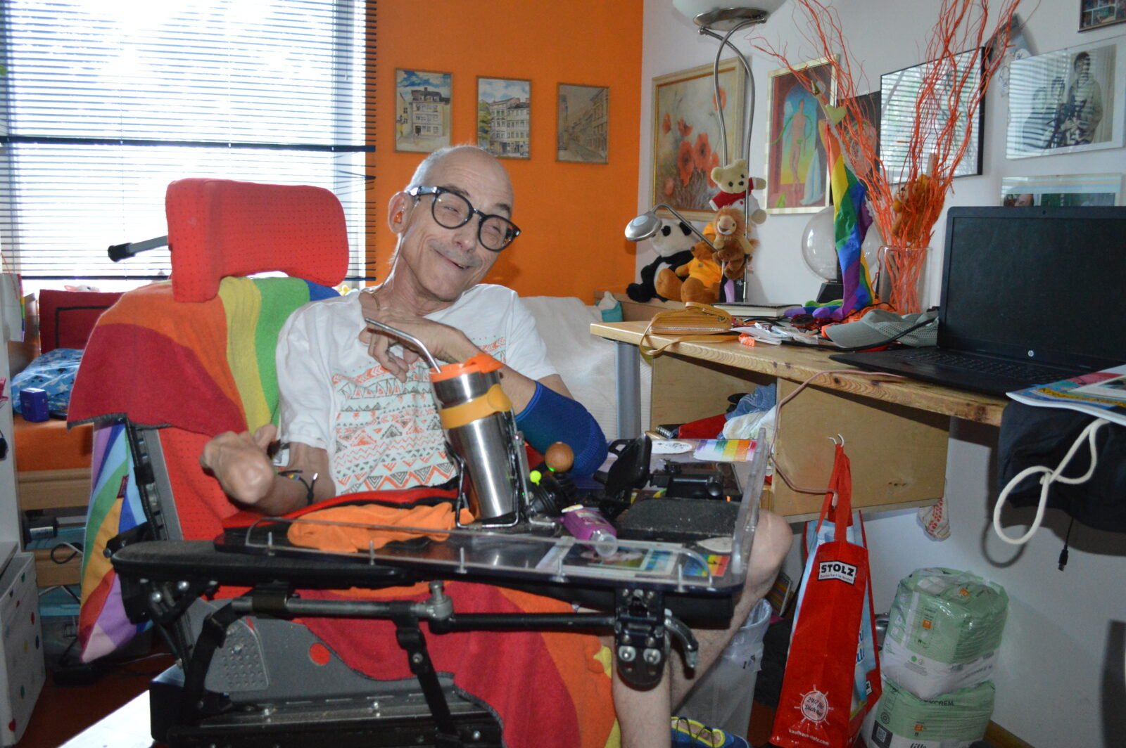 IM Bild zu sehen ist Lutz Germann, der in seinem Rollstuhl sitzt und lächelt. AM Rollstuhl und im Hintergrund sind Regenbogenflaggen.