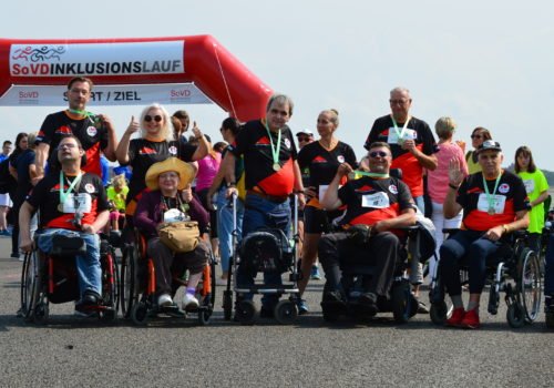 Eine Gruppe Menschen mit und ohne Rollstuhl posiert für ein Gruppenbild vor dem Start beim SoVD Inklusionslauf 2018. Alle tragen dieselben Trikots und Medaillien um den Hals.