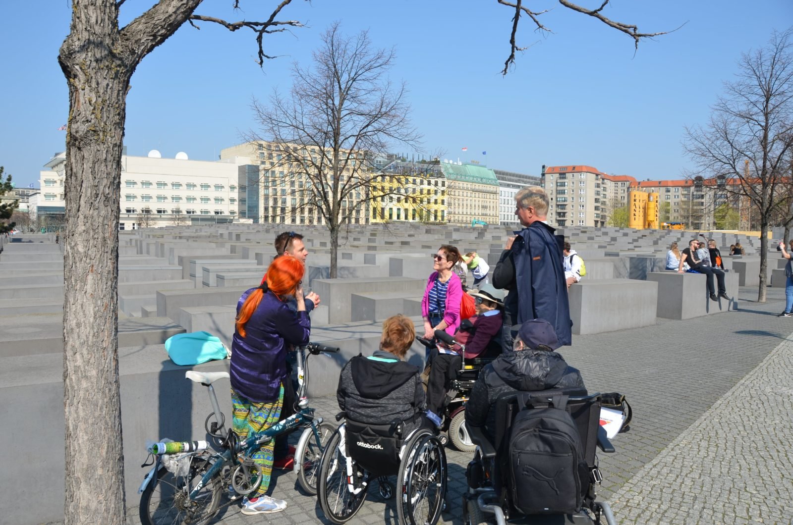 Eine Gruppe Menschen, darunter drei Rollstuhlfahrerinnen und -fahrer sowie eine Frau mit Fahrrad, steht gemeinsam vor dem Holocaust-Mahnmal in Berlin-Mitte. Der Himmel ist klar und die Sonne scheint.