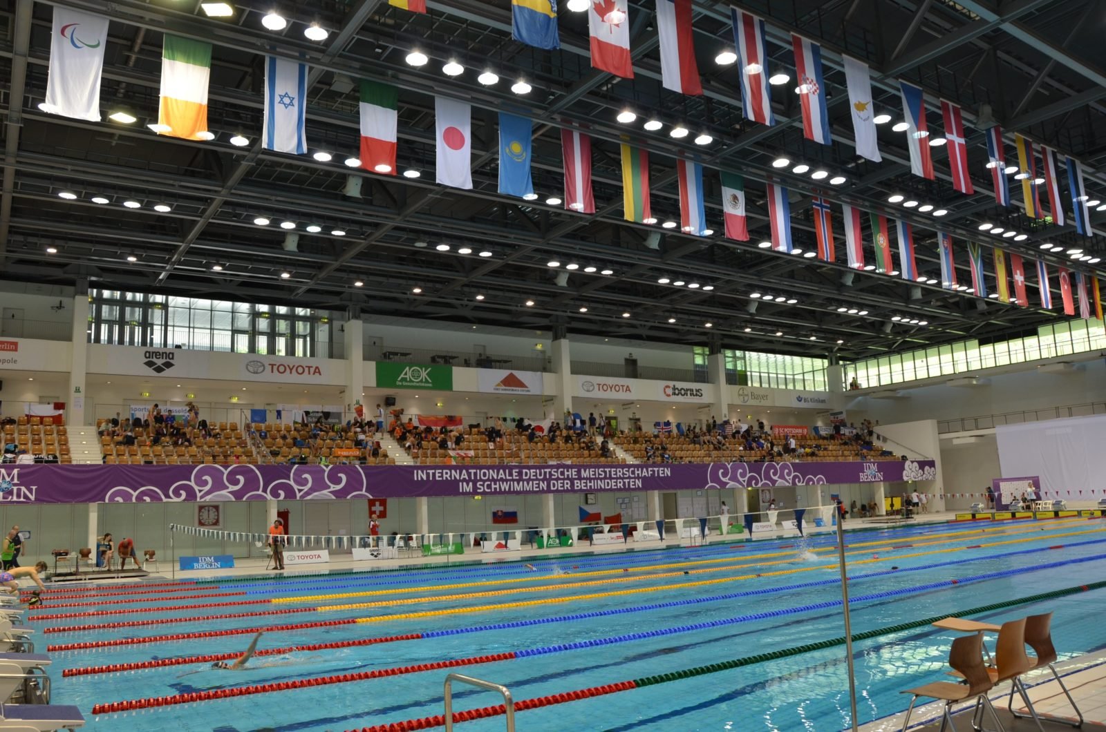 Blick in die Schwimmhalle: Vorne das Becken in dem gerade ein Wettkampf stattfindet, im Hintergrund die Ränge mit Zuschauern und darüber die Flaggen aller teilnehmenden Nationen.