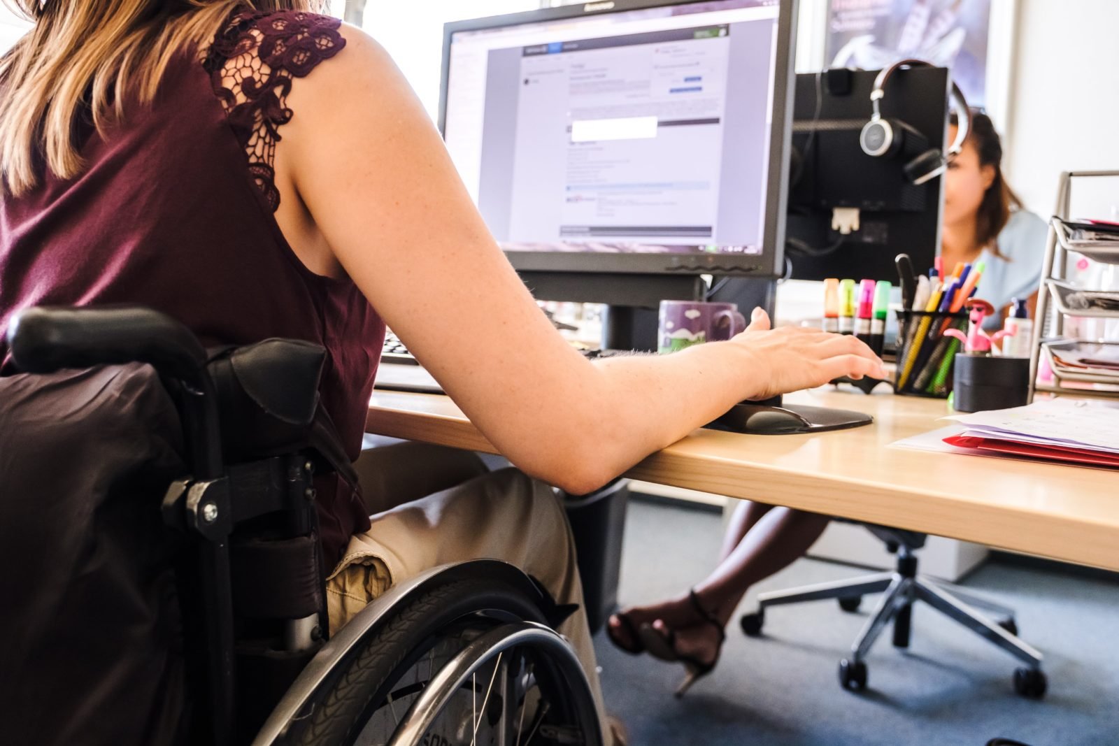 Eine junge Frau im Rollstuhl sitzt an ihrem Bildschirmarbeitsplatz. Im Hintergrund ist eine Kollegin zu sehen, die ebenfalls gerade an ihrem Bildschirm arbeitet.