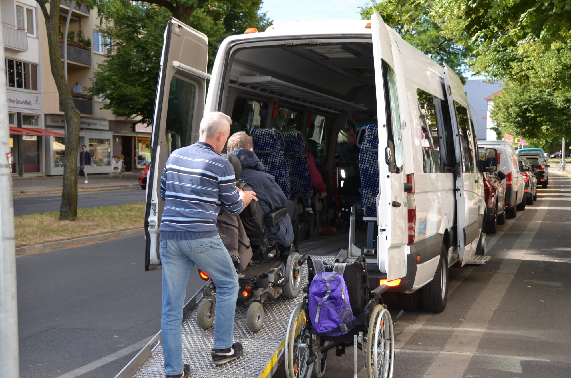 Von hinten fotografiert: ein Mann hilft einem Mann im E-Rollstuhl die Rampe in einen Sprinter-Bus hinein,