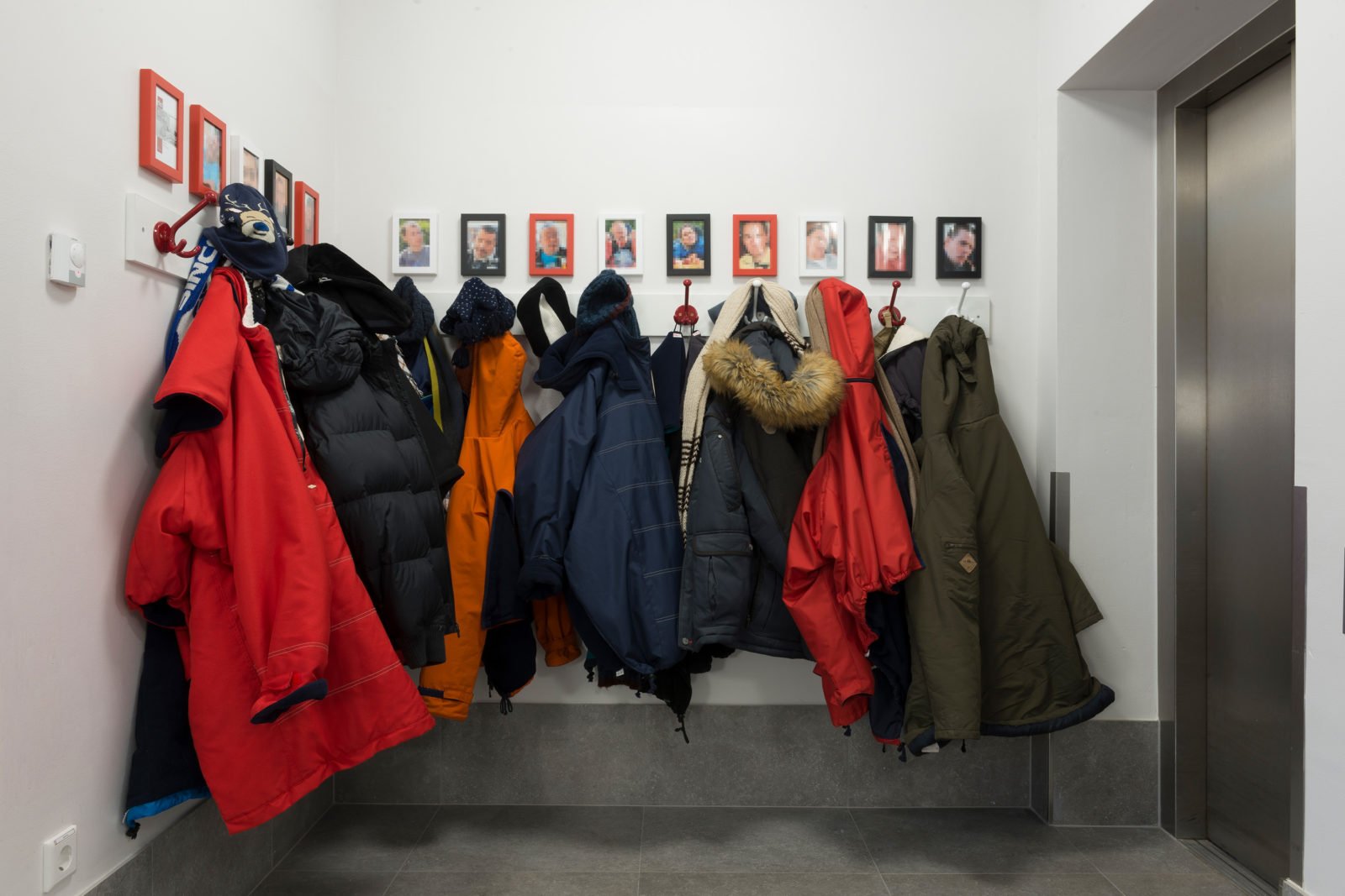 Eine Garderobe: Jacken hängen an Haken. Darüber Bilder der Bewohnerinnen und Bewohner. Diese Bilder sind unkenntlich gemacht.