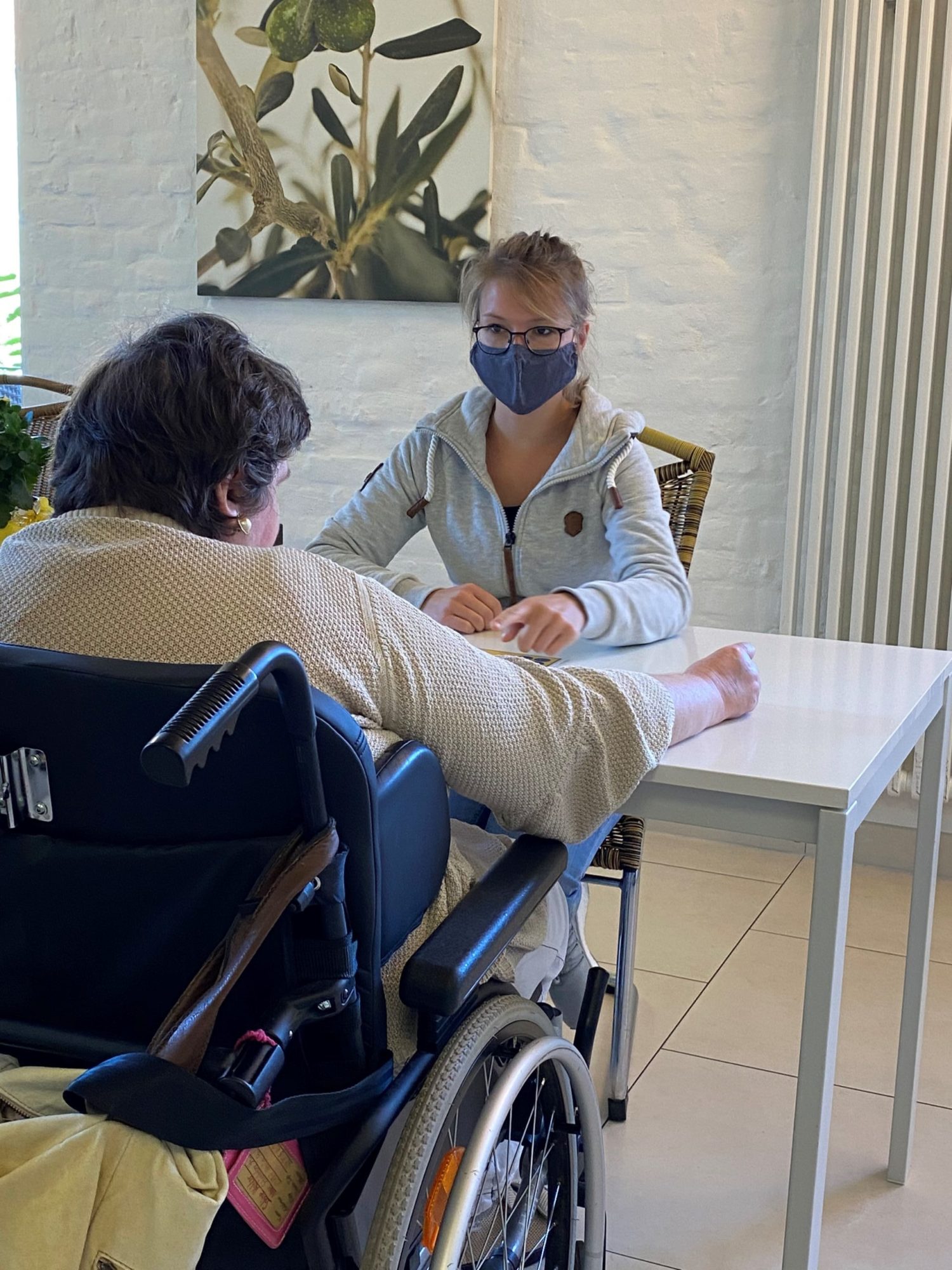 Anna Kühnert sitzt mit einer Klientin im Rollstuhl gemeinam an einem Tisch.