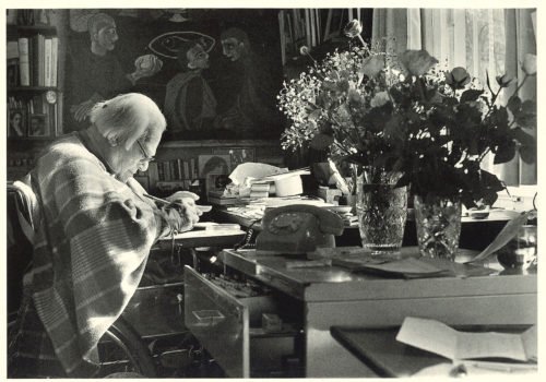 Eine Postkarte, die Lindenberg im Rollstuhl sitzen an seinem Schreibtisch zeigt. Auf demTisch stehen Blumen, ein Telefon mit Wählscheibe und diverse Dokumente und Bücher.