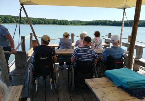 Eine Gruppe von sieben Menschen, davon drei im Rollstuhl sitzen auf einem hölzernen Floß und blicken au den See. Sie sind von hinten fotografiert, man sieht deshalb ihre Rücken. Das Wetter ist gut, das Wasser blau.