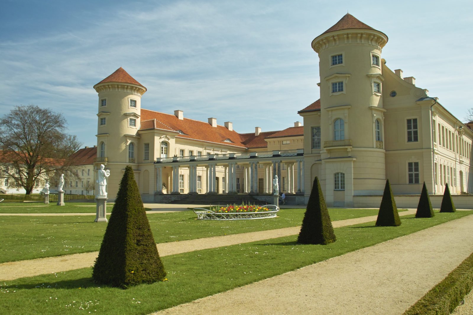Zu sehen ist das Schloss Rheinsberg von hinten. Besonders deutlich sind die beiden Türme mit einem roten Dach und einem sandfarbenen Anstrich.