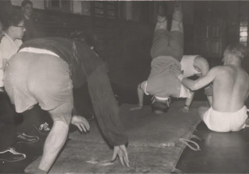 Ein altes Schwarz-Weiß-Bild aus den 60er Jahren. Es zeigt mehrere Männer, die auf Bodenmatten turnen.