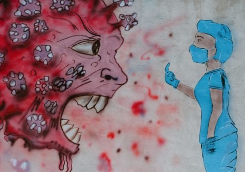 Graffiti auf Hauswand: Eine in Schutzkleidung gekleidete Person zeigt einem Cartoon-Coronavirus (mit großen Augen und scharfen Zähnen) den Mittelfinger.