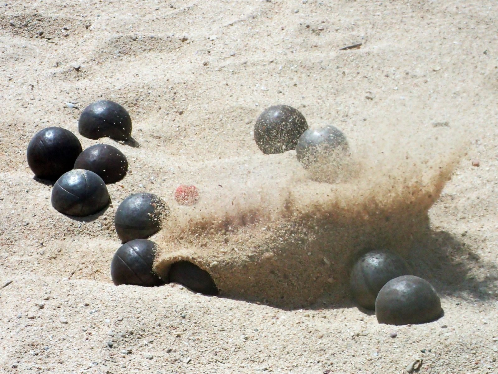 Bocciakugel schlägt im Sand ein und wirbelt ihn auf.
