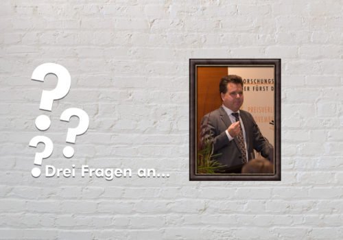 Fotomontage: Eine weiße Steinwand - darauf ein Rahmen, in den ein Bild von Jürgen Dusel montiert ist, daneben der Schriftzug Drei Fragen an... und drei Fragezeichen.