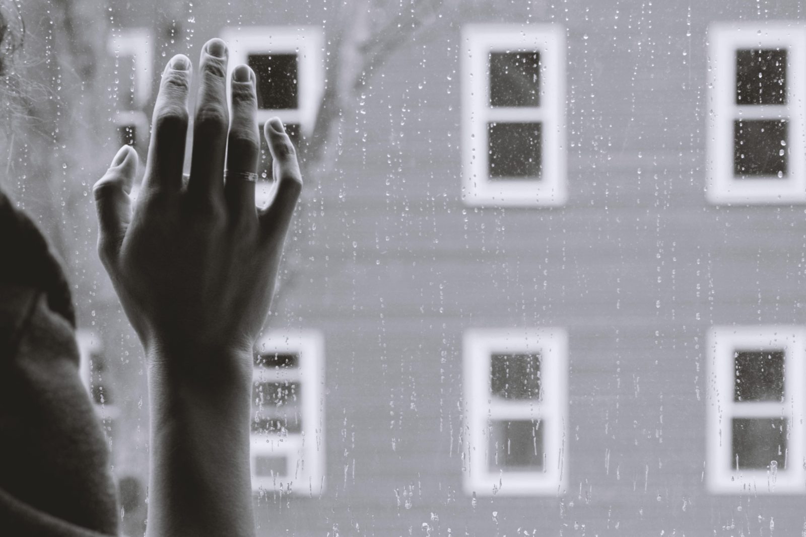 Schwarz-Weiß-Foto: Vorne eine Hand von hinten fotografiert, die an eine Fensterscheibe fasst. Im Hintergrund eine Häuserfassade mit vielen Fenstern.