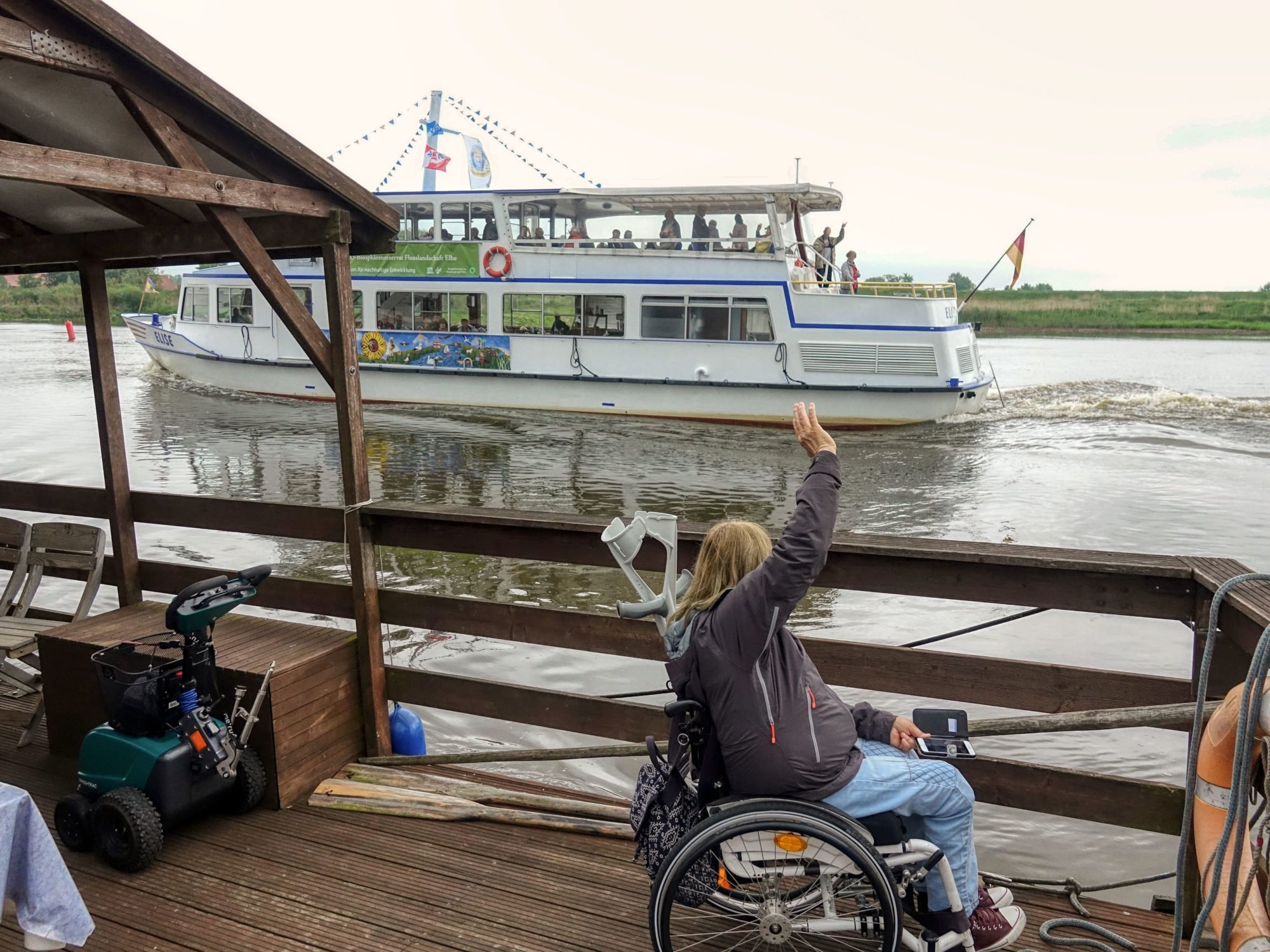 Petra Rosenberger steht mit ihrem Rollstuhl auf dem Floß und winkt den Passagieren eines vorbeifahrenden Passagierschiffes.