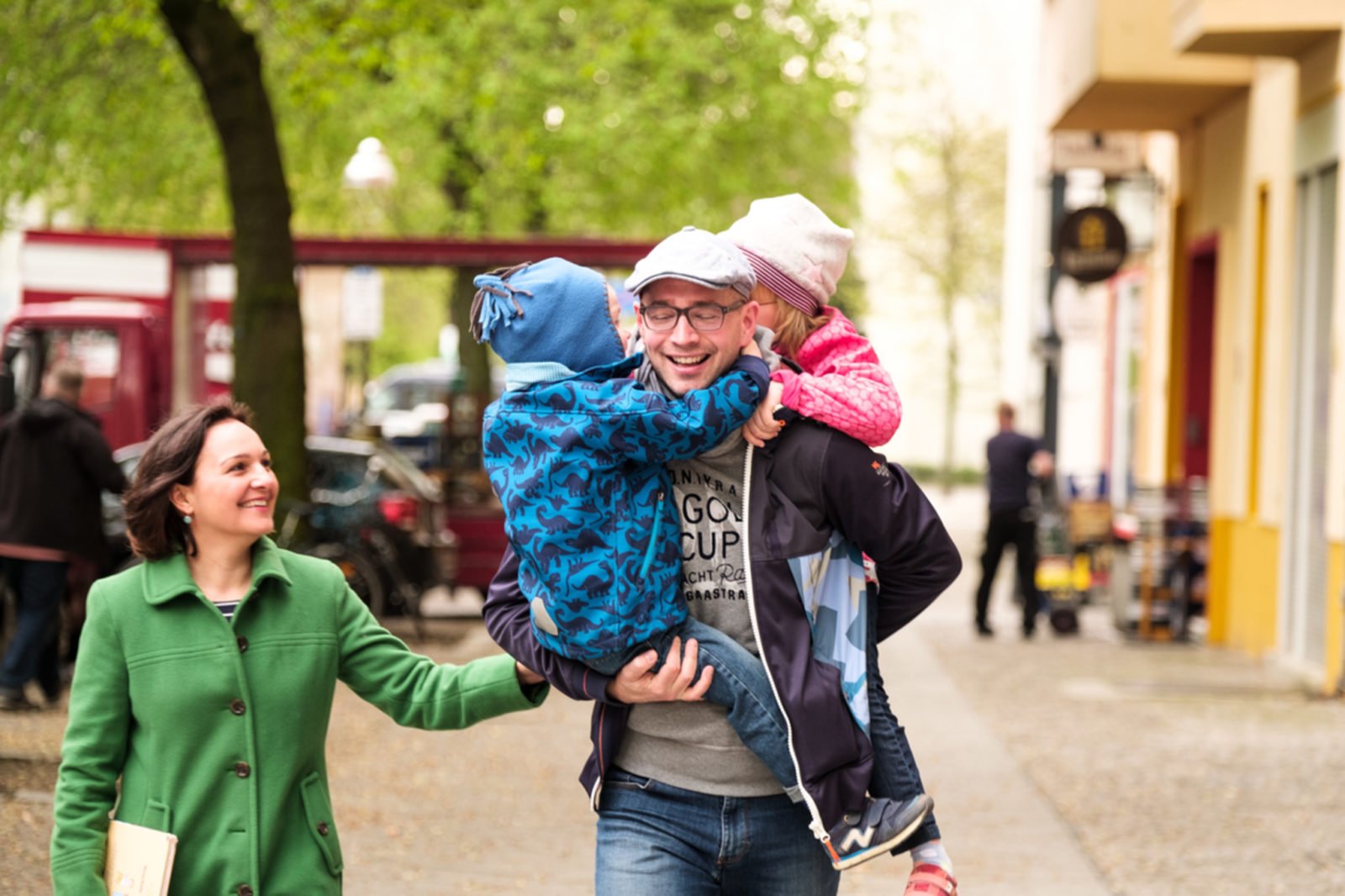 Eine vierköpfige Familie beim Spaziergang auf der Straße. Der Vater trägt seine zwei Kinder - eines auf dem Arm, das andere auf dem Rücken.