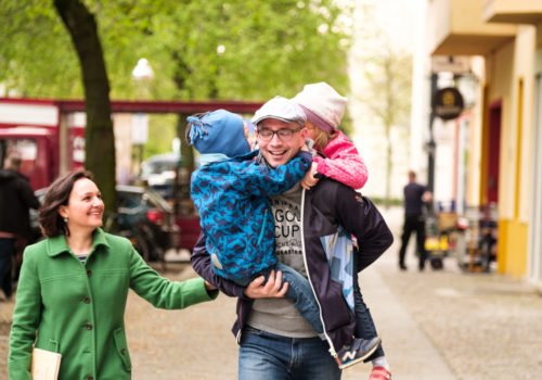 Eine vierköpfige Familie beim Spaziergang auf der Straße. Der Vater trägt seine zwei Kinder - eines auf dem Arm, das andere auf dem Rücken.