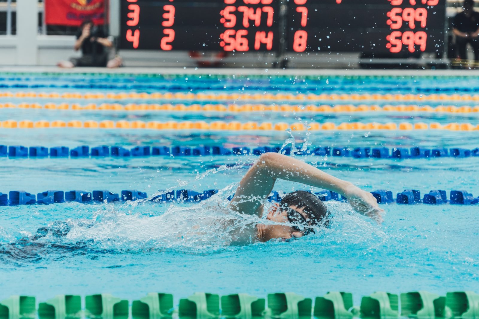 Stock-Foto: Ein junger Mann schwimmt bei einem Wettbewerb - Nahaufnahme, im Hintergrund sind Anzeigetafeln und Fotografen zu sehen.