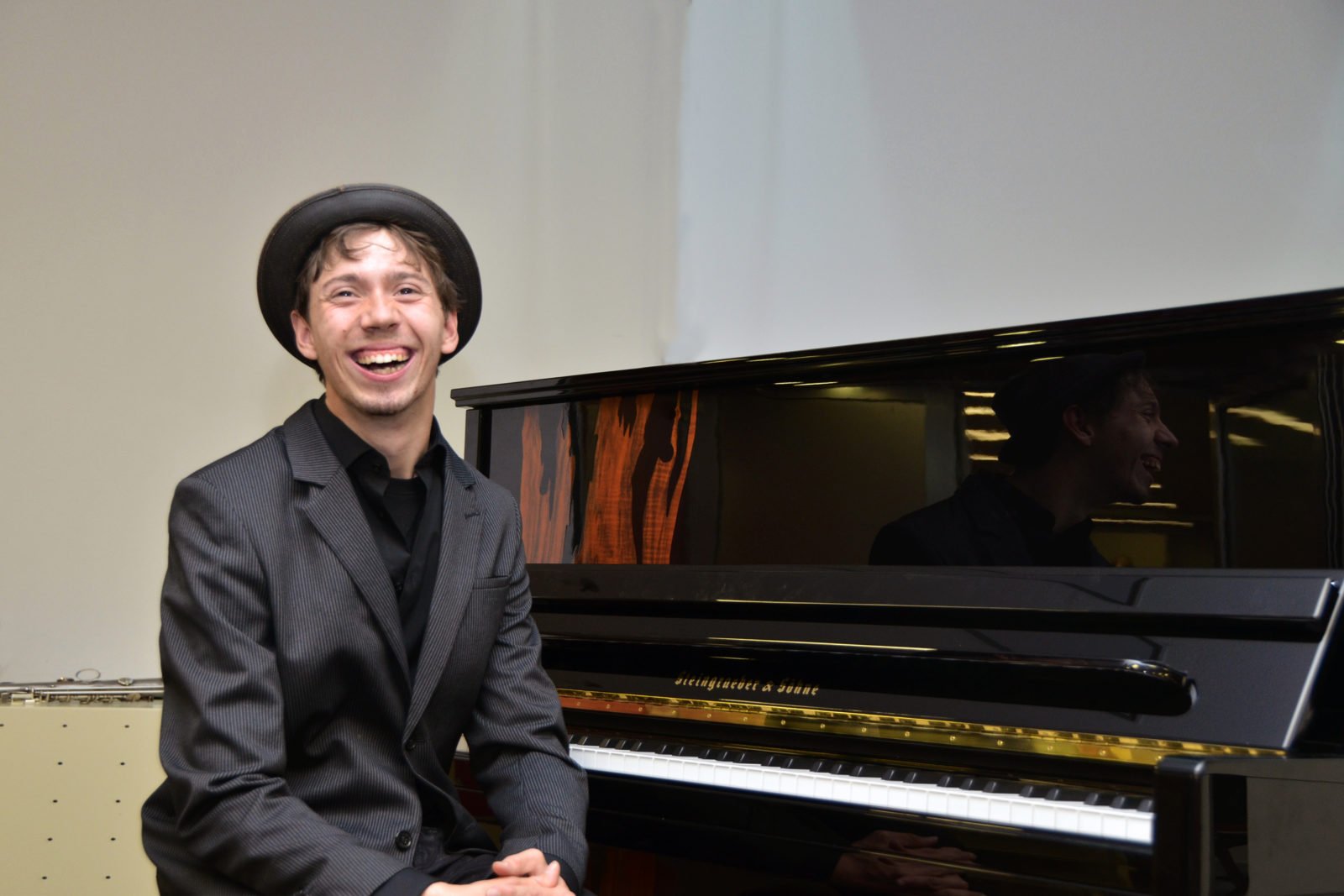 Junger Mann mit Hut sitzt vor einem Klavier und lacht in die Kamera
