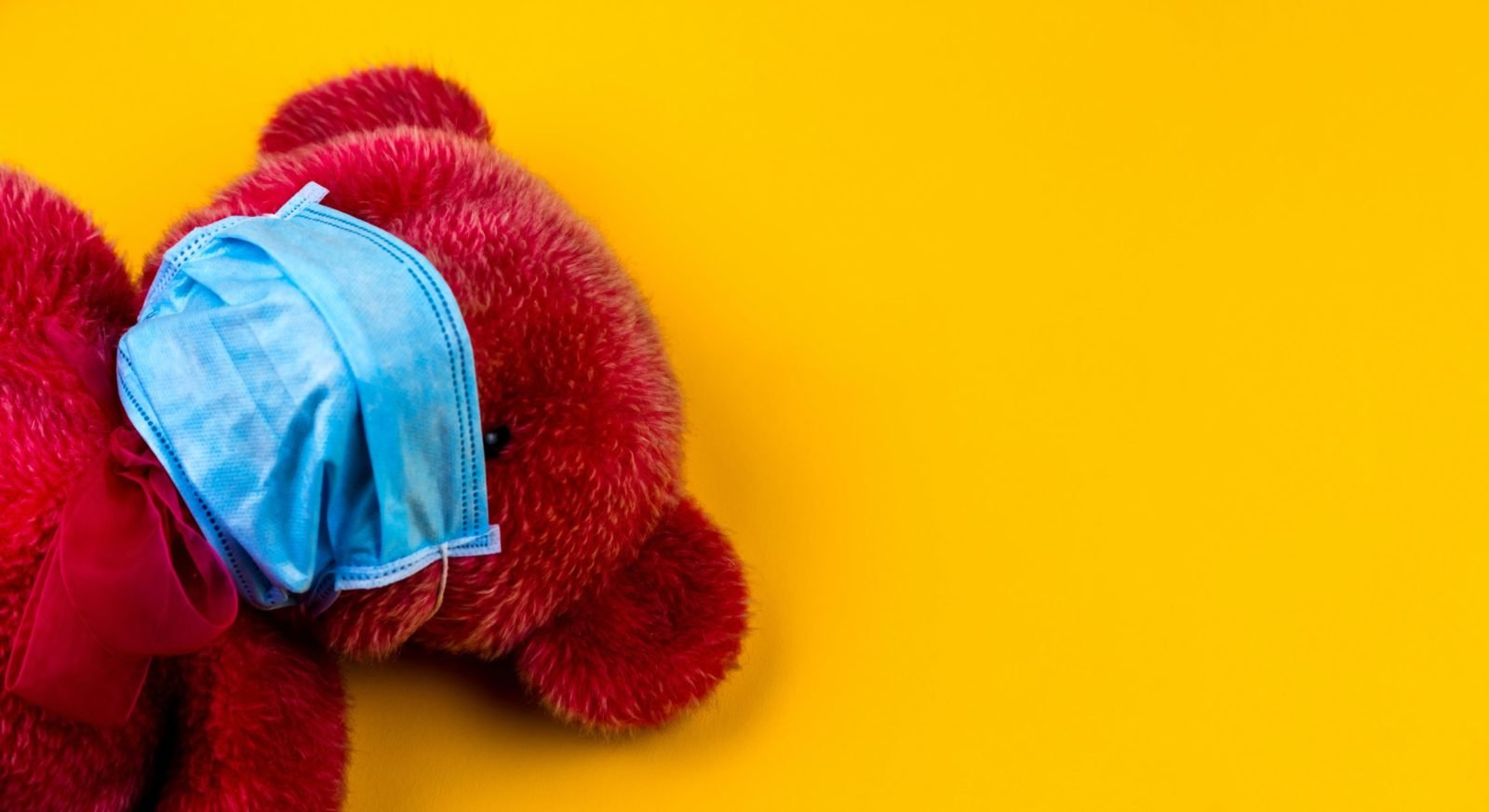 Ein roter Teddybär mit blauer OP-Maske auf gelben Hintergrund.
