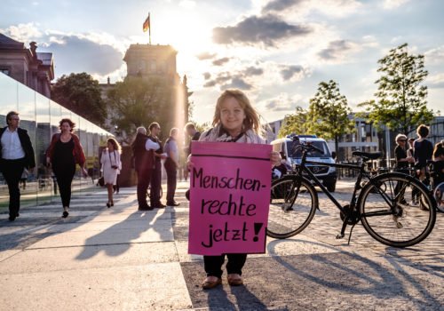 Eine Frau mit Kleinwuchs demonstriert in Berlin, vor dem Bundestag. Sie hält ein Plakat in den Händen auf dem steht: "Menschenrechte jetzt!".