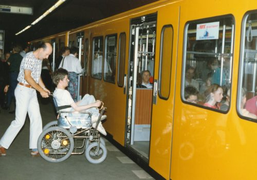 Ein altes Foto: Ein Mann hilft einem anderen Mann im Rollstuhl beim Einstieg in eine Berliner U-Bahn.