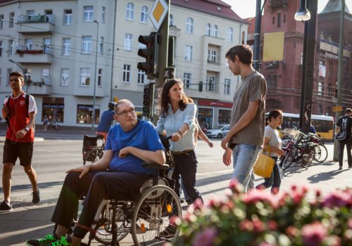 Zwei Männer, einer im Rollstuhl und einer zu Fuß, sind begleitet von einer jungen Frau in Berlin unterwegs. Alle sind gut gelaunt und lächeln.