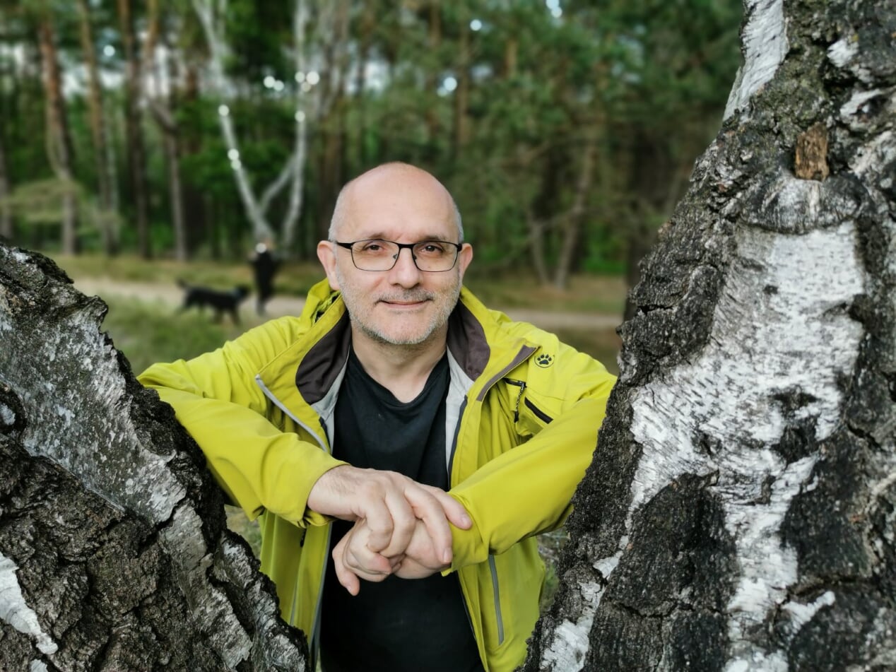 Porträtfoto von Oliver Tolmein. In Freizeitkleidung lehnt er lächelnd, zwischen zwei Baumstämmen. Im Hintergrund sind unscharf ein Waldstück, ein Weg und Fußgänger zu erkennen.