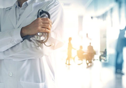 Symbolfoto: Vorne ein Arzt im Kittel mit Stethoskop. Im Hintergrund sind mehrere Menschen zu erkennen, darunter auch eine Person im Rollstuhl.