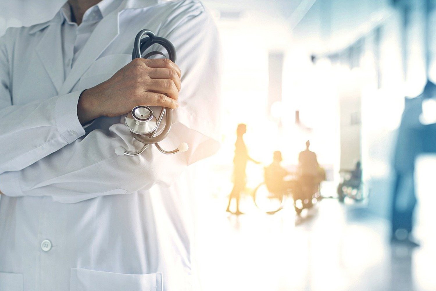 Symbolfoto: Vorne ein Arzt im Kittel mit Stethoskop. Im Hintergrund sind mehrere Menschen zu erkennen, darunter auch eine Person im Rollstuhl.